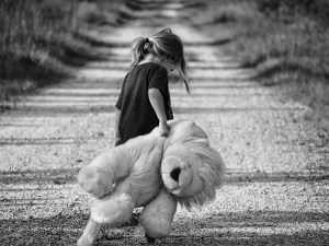 Grayscale image of a girl holding a teddy bear walking down a path with her head down