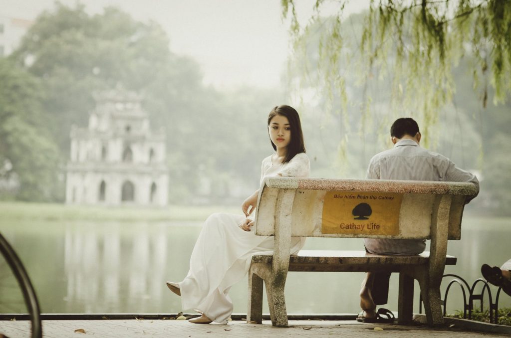 A man and woman sitting on a bench. The woman looks to the side, one leg crossed over the other with her back to the man. The man is looking down presumably reading something.