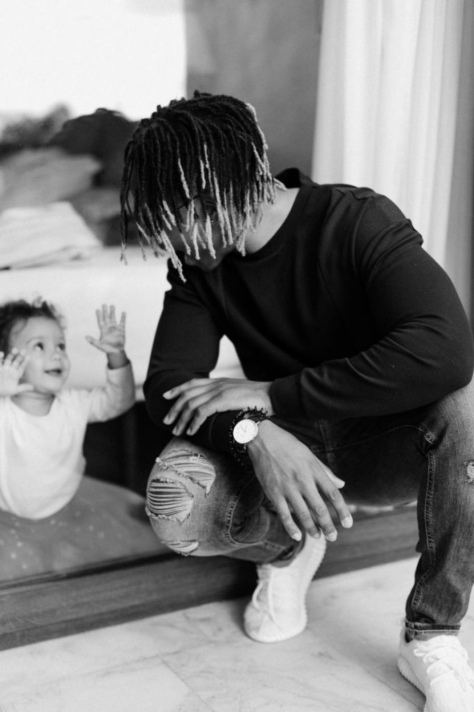 Black and white picture of black father playfully looking at his toddler through a glass door while crouching.