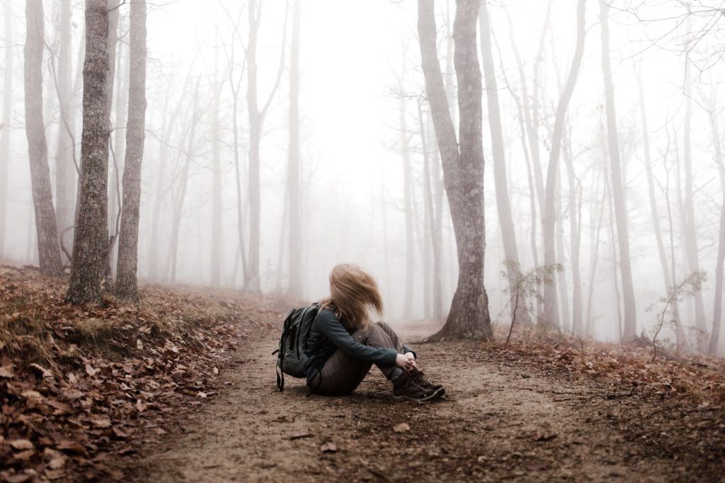 Person with hair covering their face, moving their head sitting on the ground in the woods.