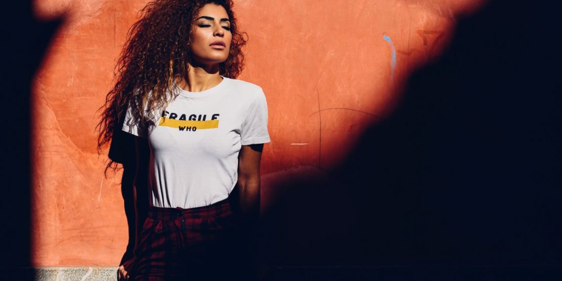 A woman of color stands resolute against a wall, her face proud, her eyes closed. Her shirt reads, "Fragile. Who?"