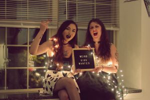 Two women holding a sign that says "Weird kids wanted" with white lights around their arms.