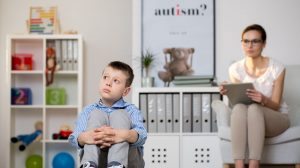 A white boy looking up and to the right looking concerned, sitting on the floor with his hands around his knees/legs, and a therapist sitting in a chair behind him staring at him with a clipboard. The poster next to the therapist says "autism?"