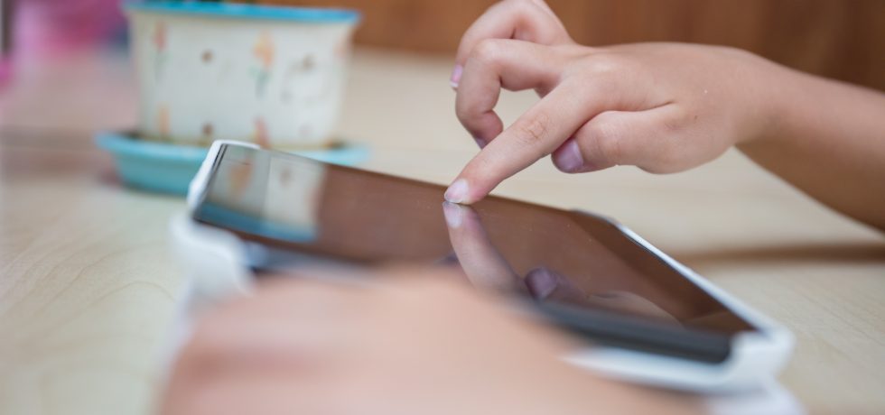 A child's hand using a tablet on a table with a desk plant in the background.