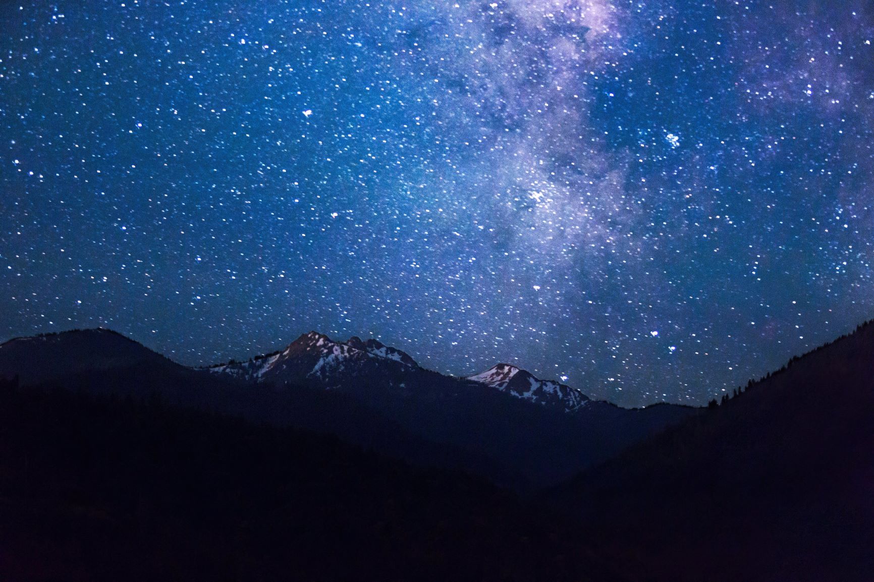 A bright, starry night sky above snow-capped forested mountains