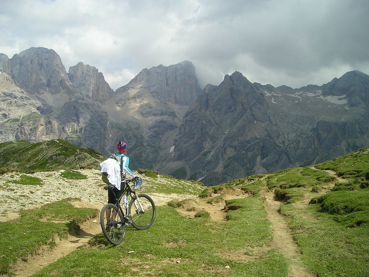 Cyclist holding a bike, standing and looking at a mountain in front of them, facing away from the camera.