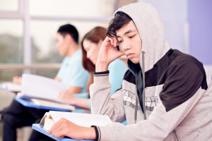 Asian teenager reading a book in a classroom with his hood up leaning on his hand.