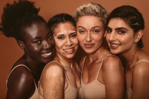 Four women with different racial backgrounds, ages, skin tones smile and stand together., and
