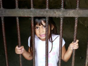 Six year old girl holds onto prison bars from inside a cell