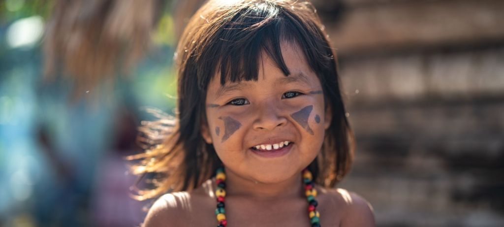 indigenous girl child about three or four years old with face paint and a beaded necklace, smiling.