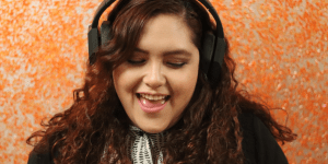 Sienna, the author of the piece, has long, curly, dark brown hair and is wearing headphones. She stands in front of an orange background